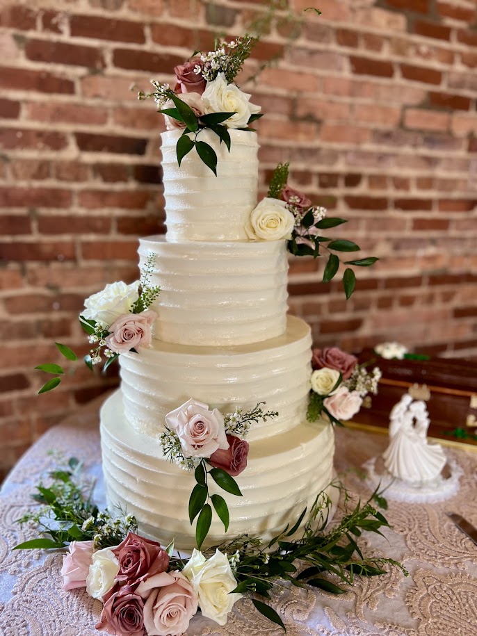 Four-tier white wedding cake decorated with blush, mauve, and ivory roses and greenery