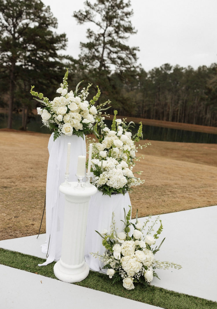 All-white ceremony florals on draped pedestals beside a candle column