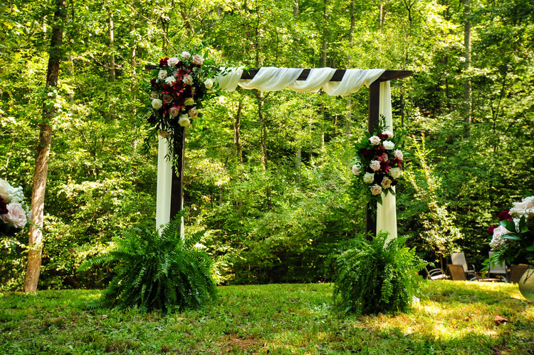 Wooden wedding arch with draped fabric and burgundy and blush rose corner florals in a forest