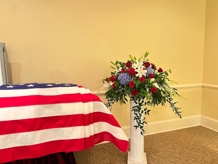Red, white, and blue pedestal arrangement of roses, lilies, and hydrangea beside a flag-draped casket