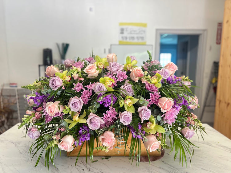 Casket spray of lavender and blush roses, green cymbidium orchids, purple stock, and palm fronds
