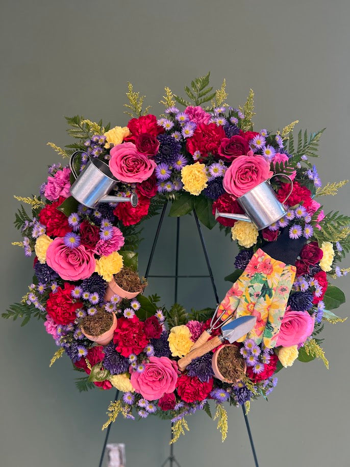 Garden-themed wreath of pink roses, red carnations, and purple asters with watering cans, gardening gloves, and a trowel